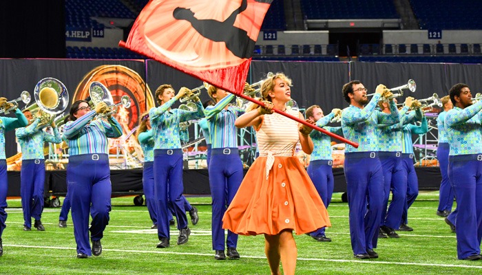 Color guard performer twirls a flag while marching band members play brass instruments on a stadium field