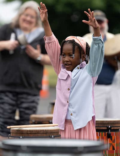 Young girl smiles and raises both arms joyfully during an outdoor music activity