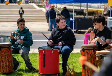 Three young boys sit on grass playing red conga drums together outdoors