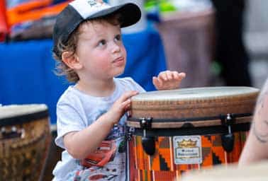 A toddler in a cap curiously touches a conga drum at a community event kid zone