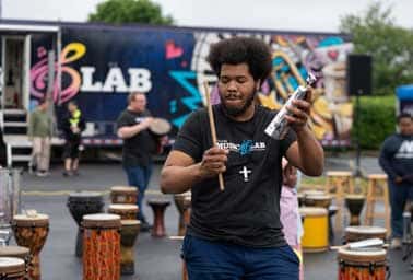A man plays a djembe drum while walking near the Mobile Music Lab van at an outdoor event
