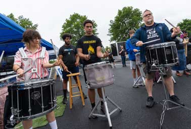 Three men sit on stools playing snare drums together under a tent outdoors