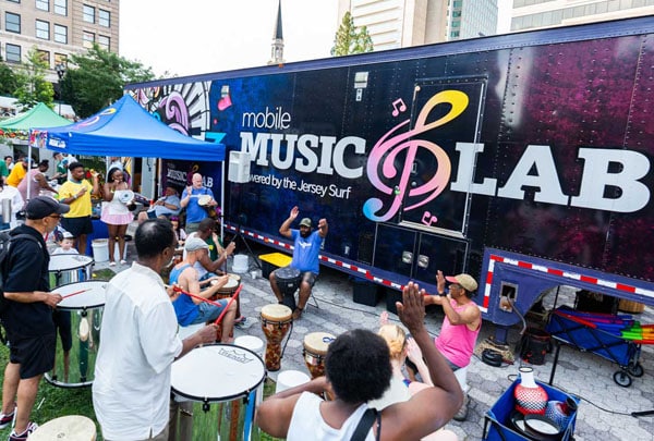 Community members play djembe drums in a street festival drum circle next to the Mobile Music Lab trailer