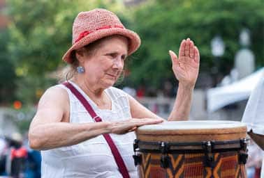 A woman in a red hat raises her hand while playing a djembe drum outdoors