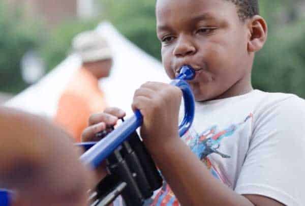 A boy blows a blue plastic horn as another child's hand appears in the foreground outdoors