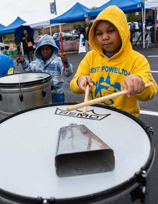 A boy in a yellow hoodie plays a marching snare drum outdoors with drumsticks
