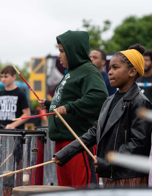 Students in hoodies watch attentively while one holds a drumstick during an outdoor school event
