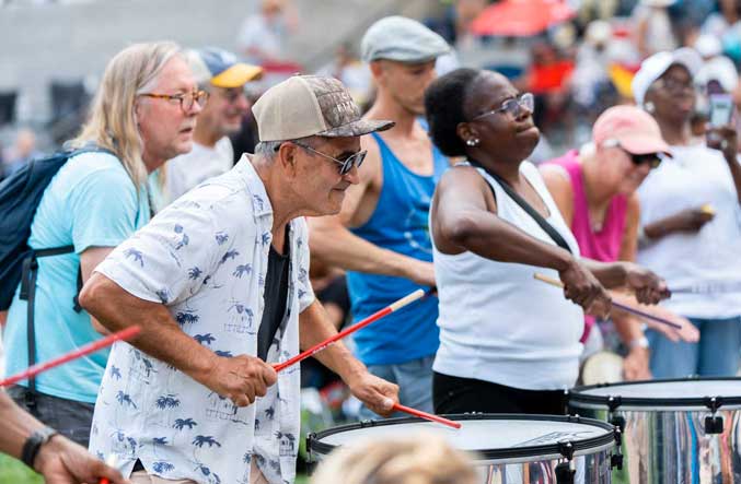 A row of older adults plays floor drums with sticks at an outdoor wellness event