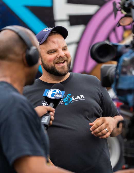 A bearded man wearing headphones smiles while covering a Drum Jawn event for the news