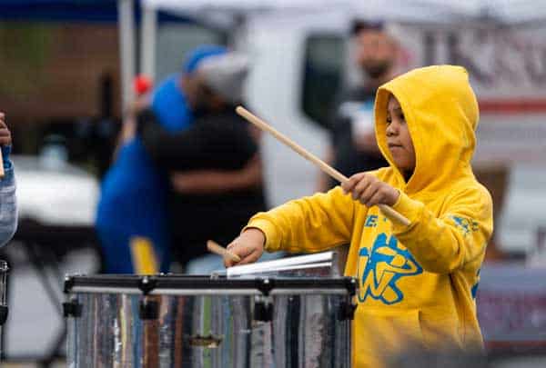 A young drummer in a yellow hoodie raises a drumstick mid-beat during a school program performance