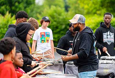 An instructor guides a group of teenage students playing tenor drums outdoors
