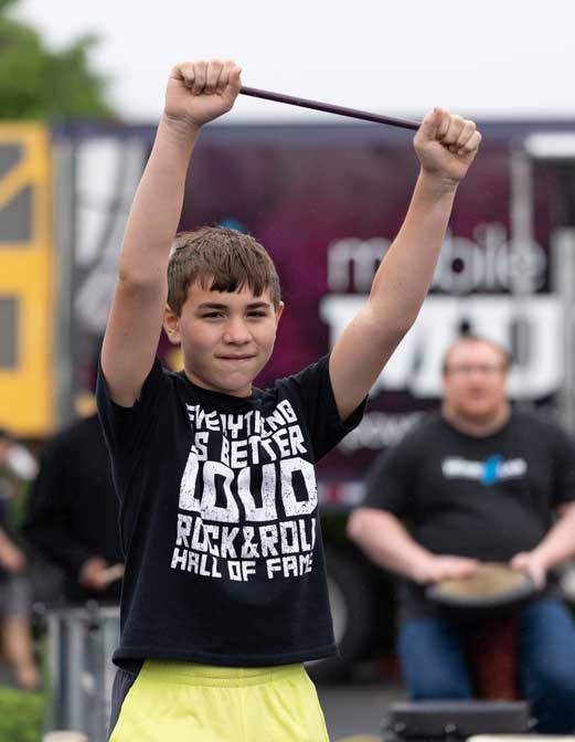 A smiling boy raises a drumstick overhead with both hands outdoors