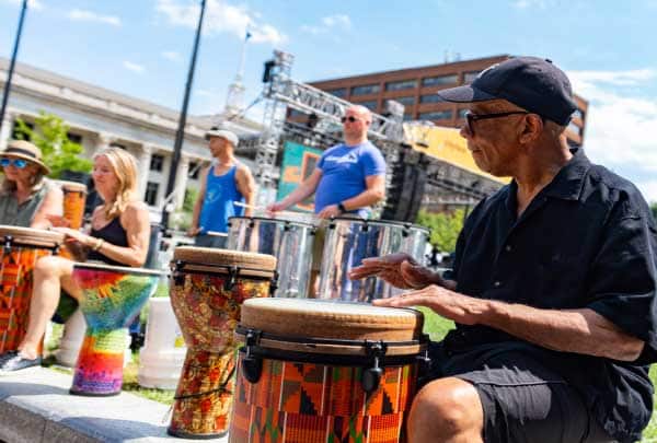 A row of adults plays djembe drums together outdoors in an urban drum circle