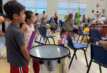 Children stand near a large drum in a classroom during a small group music lesson