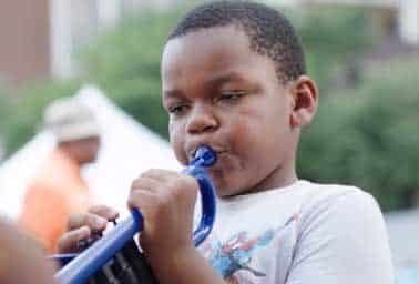 Young boy blows into a blue plastic horn while an adult in orange looks on in the background