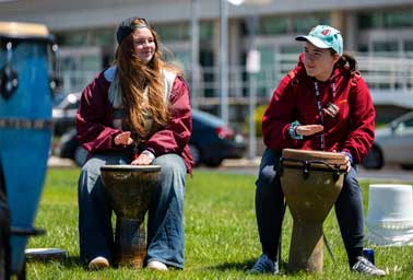 Two teens sit outdoors playing djembe drums during an instrument discovery session