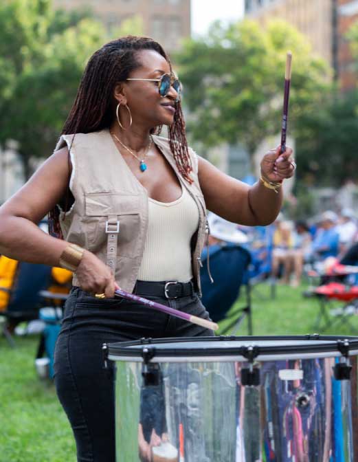 A woman plays a floor drum with sticks at an outdoor community music event